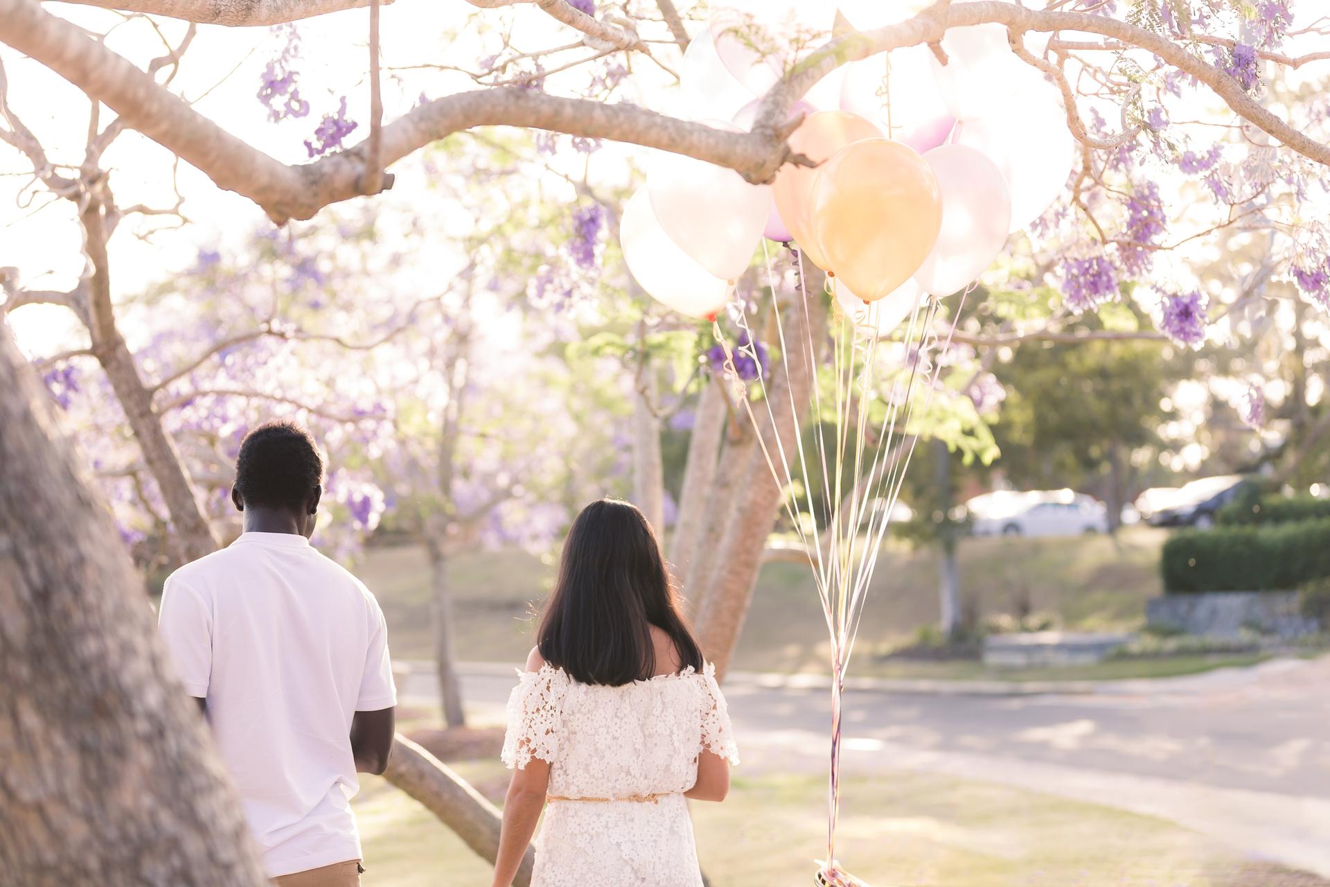 The couple walking under jacaranda trees at UQ Lakes, holding colorful balloons, with soft sunlight filtering through.