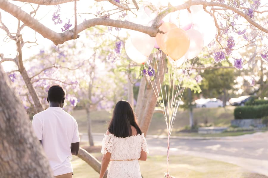The couple walking under jacaranda trees at UQ Lakes, holding colorful balloons, with soft sunlight filtering through.