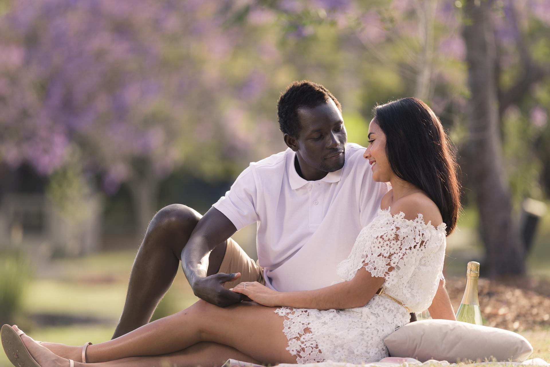 The couple sitting on a blanket at UQ Lakes, gazing at each other affectionately, surrounded by blooming jacarandas.