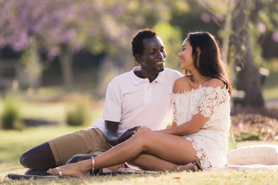 The couple sitting close on a picnic blanket, smiling warmly at each other, with jacaranda blossoms in the background.
