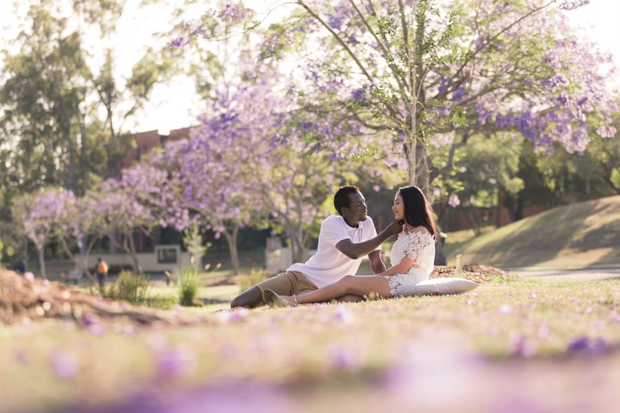 The couple sitting on the grass, laughing together, with a backdrop of purple jacaranda flowers.
