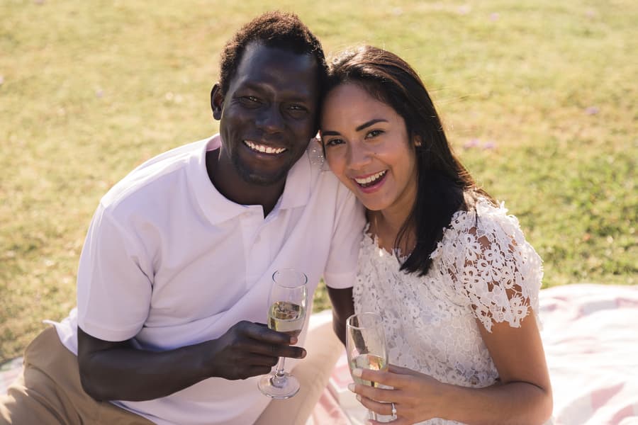 The couple sitting on a blanket, holding glasses of champagne, smiling at the camera with jacarandas in the background.