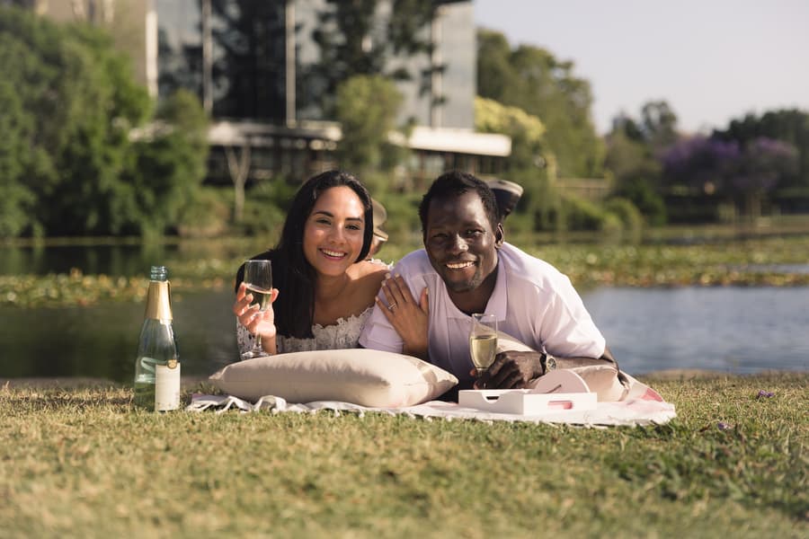 The couple lying on a blanket by the lake, holding champagne glasses, smiling at the camera.