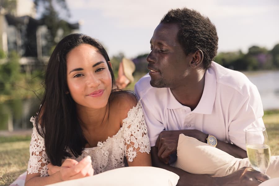 The couple lying on grass at UQ Lakes, smiling warmly at the camera in soft afternoon light.