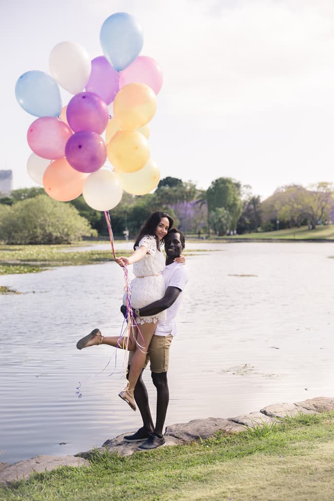 One partner holding colorful balloons while being lifted by the other near the lake.