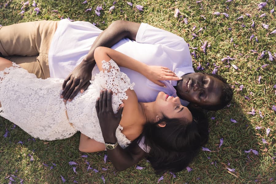 The couple lying on grass surrounded by purple petals, embracing and smiling peacefully.