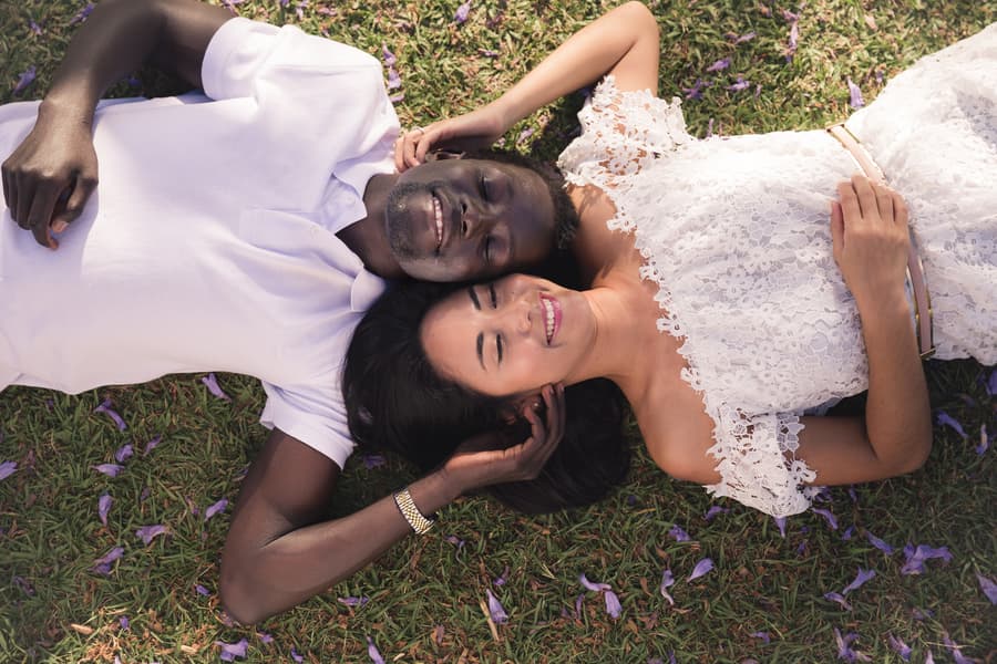 The couple lying on grass, heads together, smiling up at the sky with closed eyes.