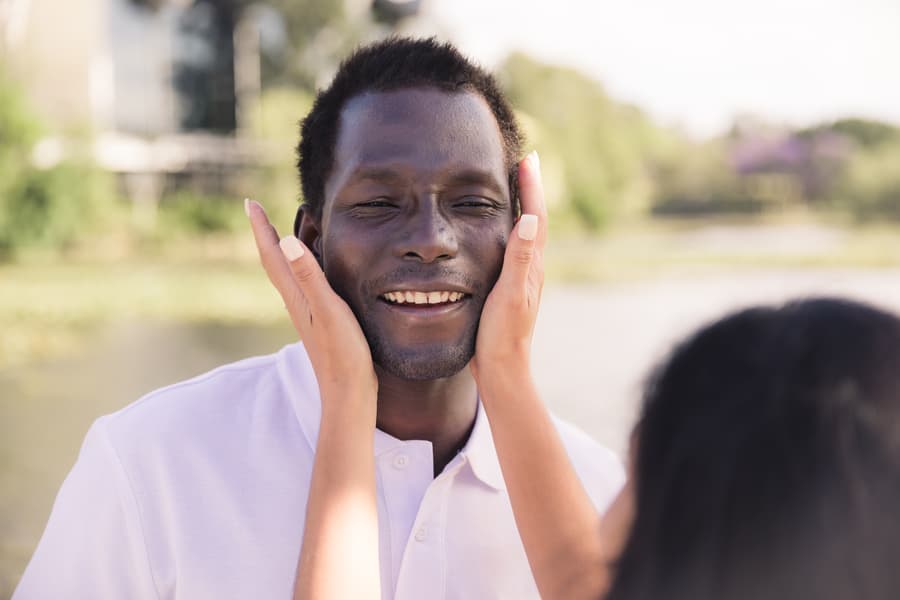 One partner gently holding the other's face, both smiling warmly by the lake.
