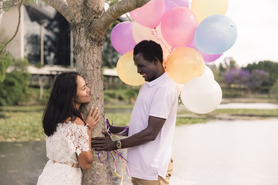 The couple standing by a tree with colorful balloons, gazing at each other affectionately.