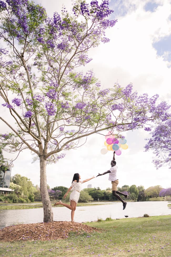 One partner holding balloons, playfully jumping while the other reaches out by a tree.