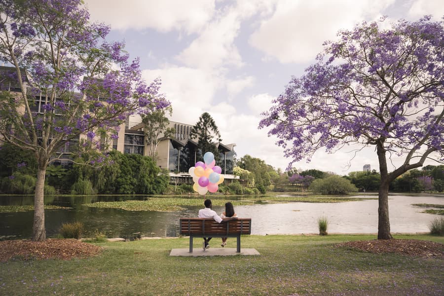 The couple sitting on a bench by the lake, surrounded by purple-flowered trees, holding balloons.