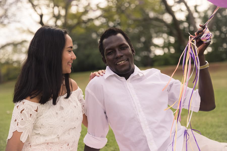 The couple sitting on grass, one holding balloons, both smiling at each other warmly.