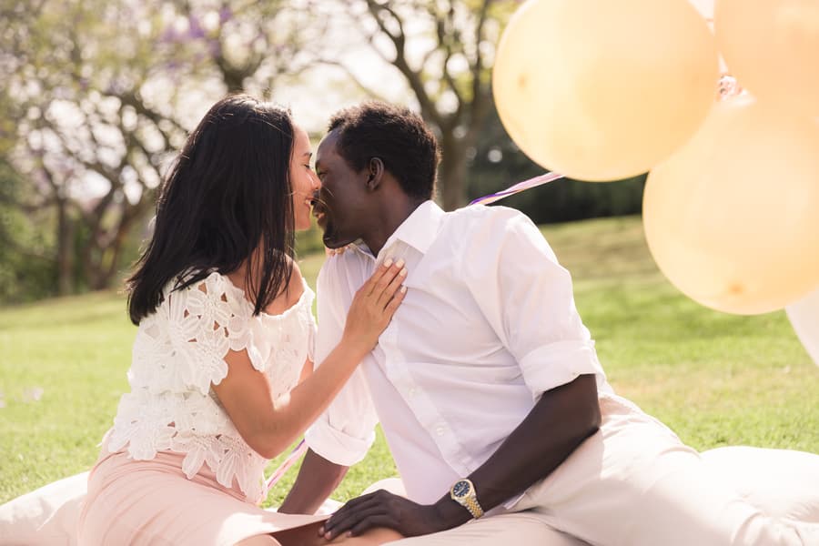 The couple sitting closely on grass, leaning in for a kiss, surrounded by balloons.