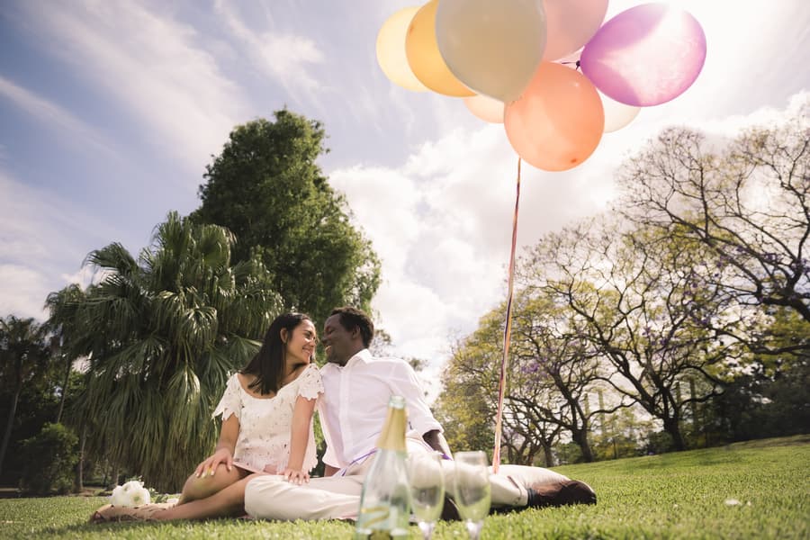 A couple sitting on grass at UQ Lakes, surrounded by trees and holding colorful balloons, with a bottle of champagne nearby.