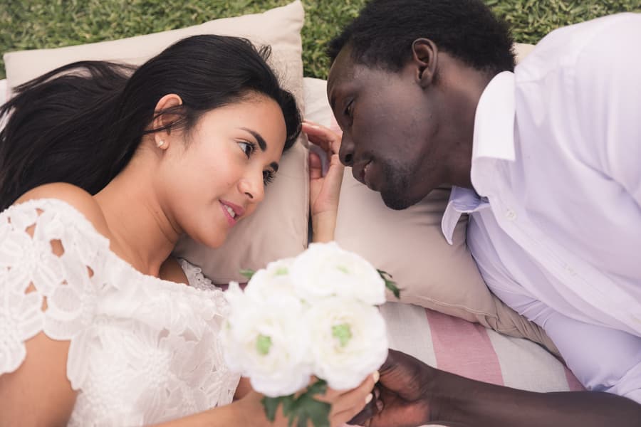 A couple lying on a blanket on grass, gazing into each other's eyes, with the woman holding a bouquet of white flowers.