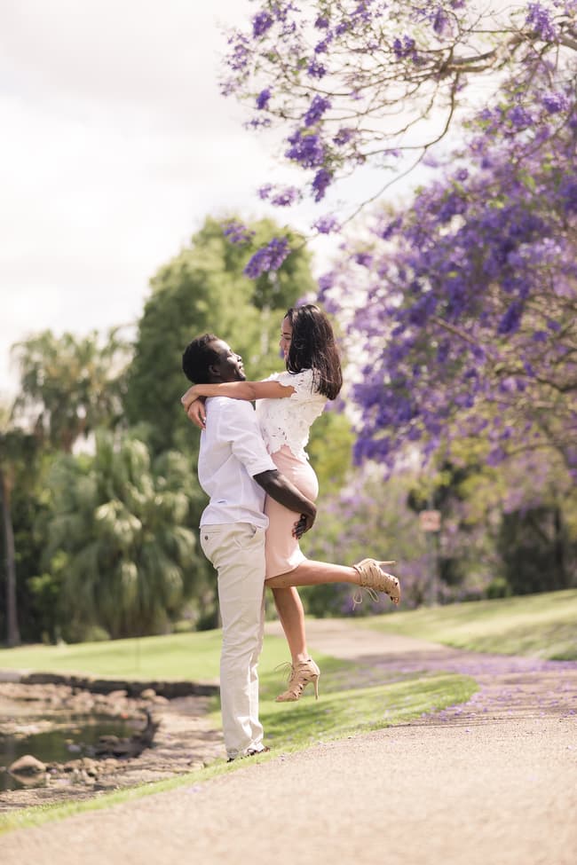 A man lifting a woman joyfully under jacaranda trees, with purple flowers scattered on the ground.