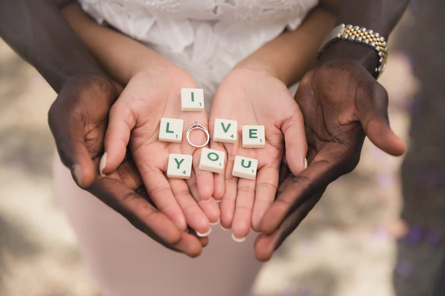 Hands of a couple holding Scrabble tiles spelling 'I LOVE YOU' with an engagement ring placed on the tiles.