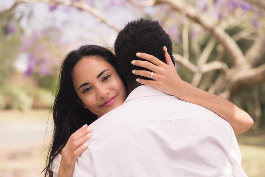 A woman embracing a man, showing an engagement ring on her hand, with a content expression.