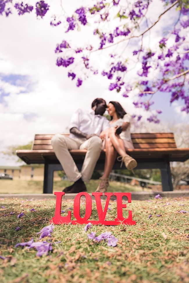 A couple sitting on a bench under jacaranda trees, with a red 'LOVE' sign on the grass in the foreground.