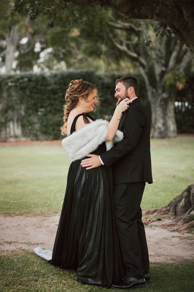 The bride Jayme and groom Christopher embrace outdoors under a large tree, with Jayme wearing a black dress and white shawl and Christopher in a black suit. Award winning wedding photographer by She Said Yes.