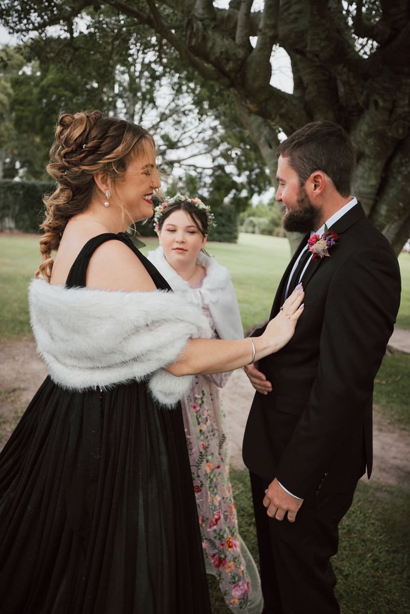 The bride Jayme in a black dress and white fur shawl touches the groom Christopher's chest while a bridesmaid in a floral dress and white shawl stands behind them outdoors. Candid wedding photographer by She Said Yes.