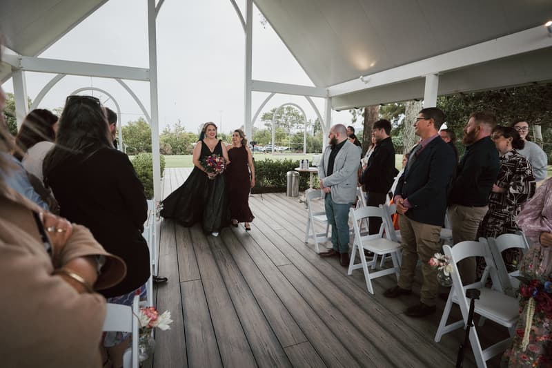 The bride Jayme, wearing a black dress and holding a bouquet, walks down the aisle at Sandstone Point Hotel — The Pavilion accompanied by a woman in a dark dress, while guests stand on either side watching.