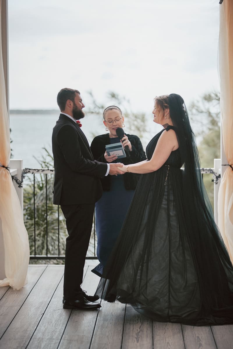 Bride Jayme and groom Christopher hold hands facing each other during their wedding ceremony, with the officiant reading vows between them. Spring wedding Queensland venues by She Said Yes.