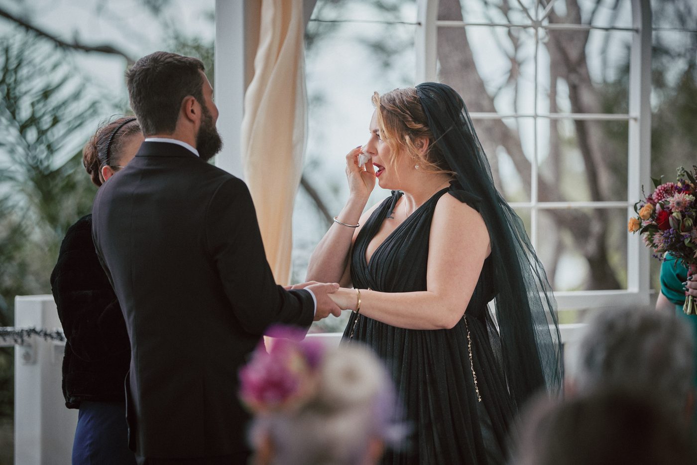 Bride Jayme in a black dress and veil wipes her eye while holding hands with groom Christopher during their wedding ceremony. Destination wedding video packages by She Said Yes.
