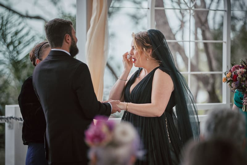 Bride Jayme in a black dress and veil wipes her eye while holding hands with groom Christopher during their wedding ceremony. Destination wedding video packages by She Said Yes.