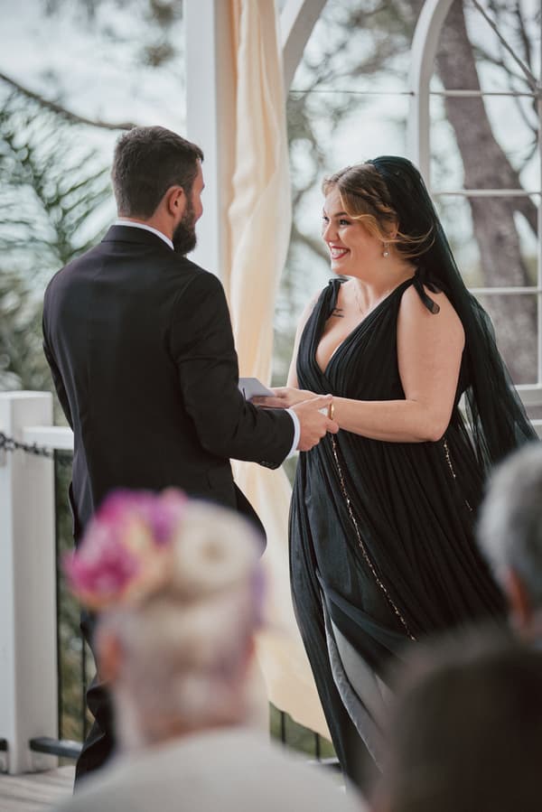 Jayme and Christopher hold hands facing each other during their wedding ceremony at Sandstone Point Hotel — The Pavilion, with guests visible in the foreground.