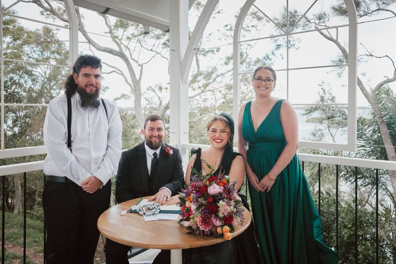Bride Jayme and groom Christopher sit at a round wooden table with a large floral bouquet, signing documents at Sandstone Point Hotel — The Pavilion, accompanied by a bridesmaid in a green dress and a groomsman in a white shirt with suspenders.