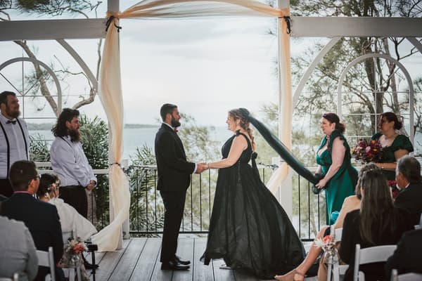 Bride Jayme and groom Christopher hold hands facing each other during their wedding ceremony, with bridesmaids and groomsmen standing nearby and guests seated watching. Event videographer Brisbane by She Said Yes.