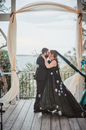 Bride Jayme and groom Christopher share a kiss at the ceremony stage under a decorated arch at Sandstone Point Hotel — The Pavilion, with bubbles floating around them and a waterfront view in the background.