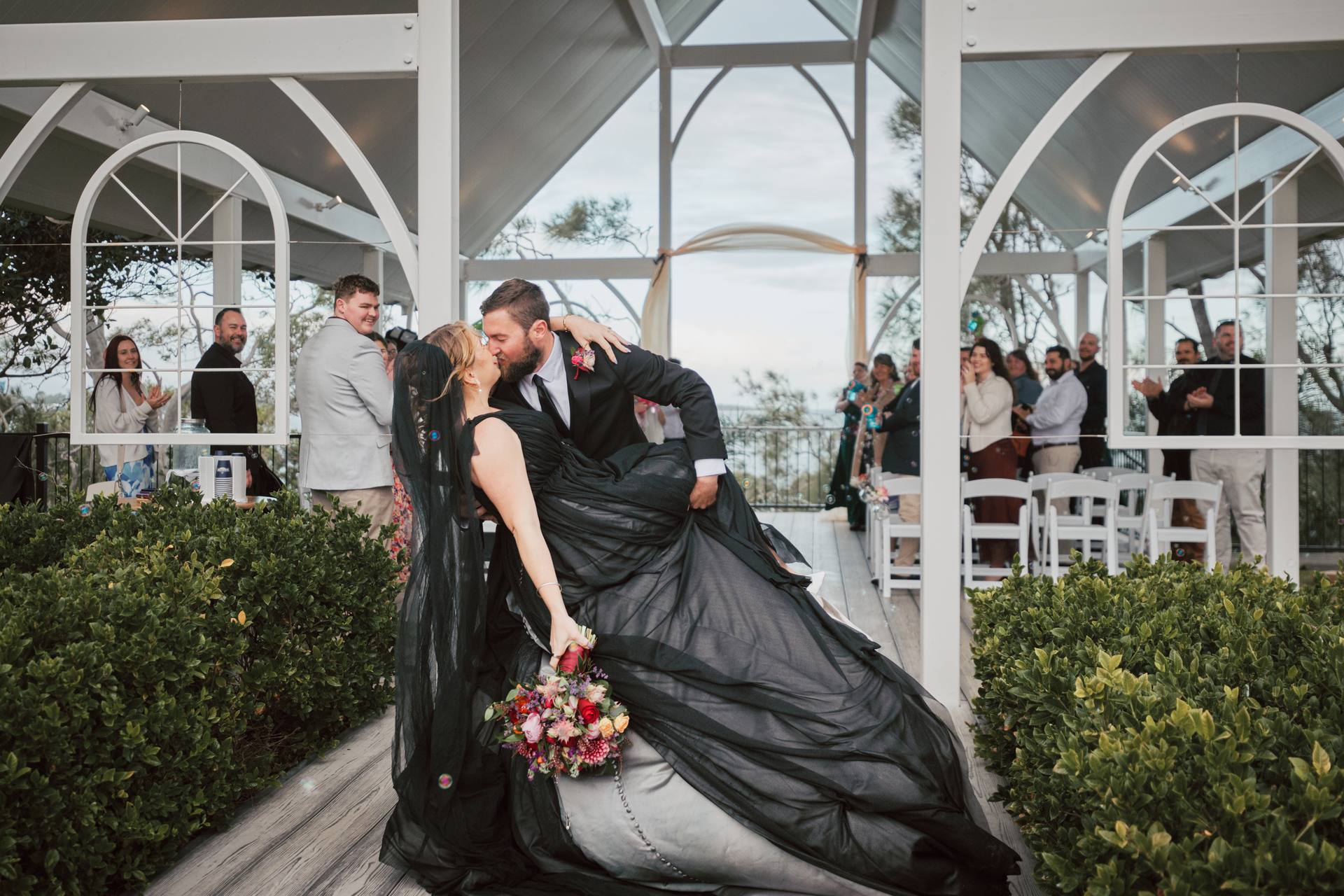 Bride Jayme in a black wedding dress and groom Christopher share a kiss as he dips her at the ceremony stage at Sandstone Point Hotel — The Pavilion, with guests applauding in the background.