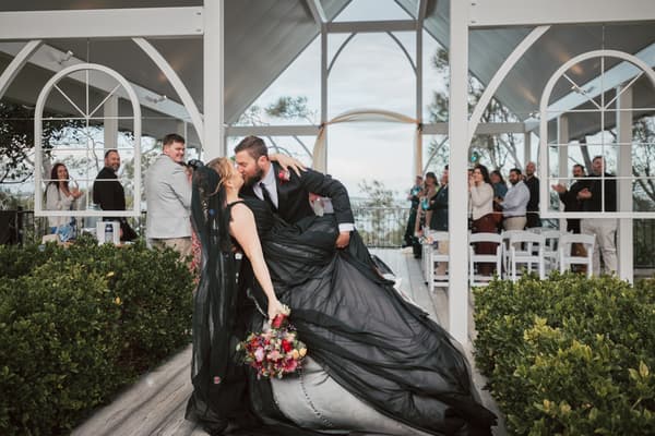 Bride Jayme in a black wedding dress and groom Christopher share a kiss as he dips her at the ceremony stage at Sandstone Point Hotel — The Pavilion, with guests applauding in the background.