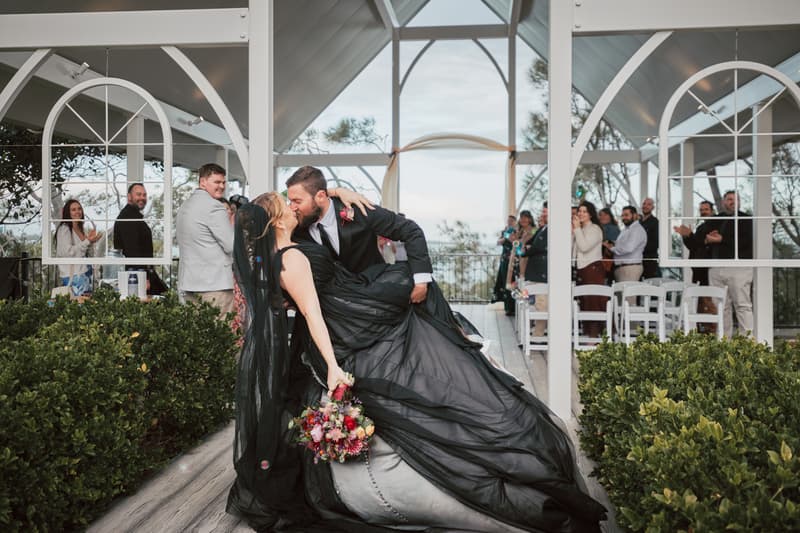 Bride Jayme in a black wedding dress and groom Christopher share a kiss as he dips her at the ceremony stage at Sandstone Point Hotel — The Pavilion, with guests applauding in the background.