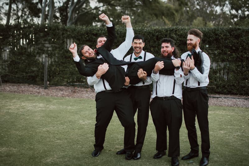 Christopher and four groomsmen pose outdoors on grass at Sandstone Point Hotel — The Pavilion, with the groomsmen holding Christopher horizontally.