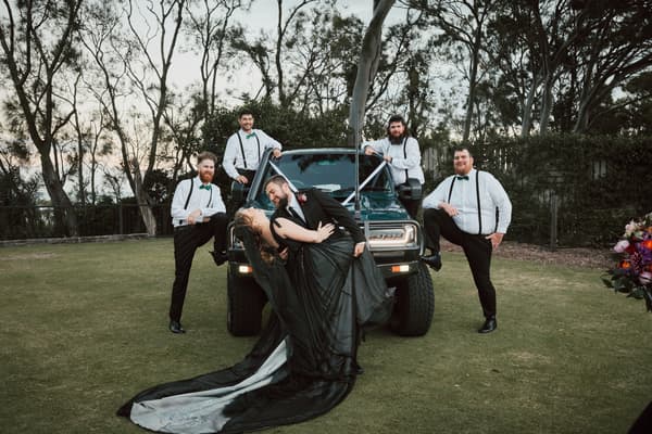 The bride and groom pose in front of a green vehicle outdoors with four groomsmen standing around them, all dressed in white shirts with black suspenders and green bow ties. Best wedding photographer Moreton Bay by She Said Yes.