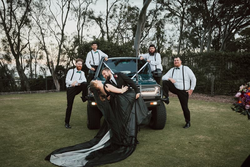 The bride and groom pose in front of a green vehicle outdoors with four groomsmen standing around them, all dressed in white shirts with black suspenders and green bow ties.