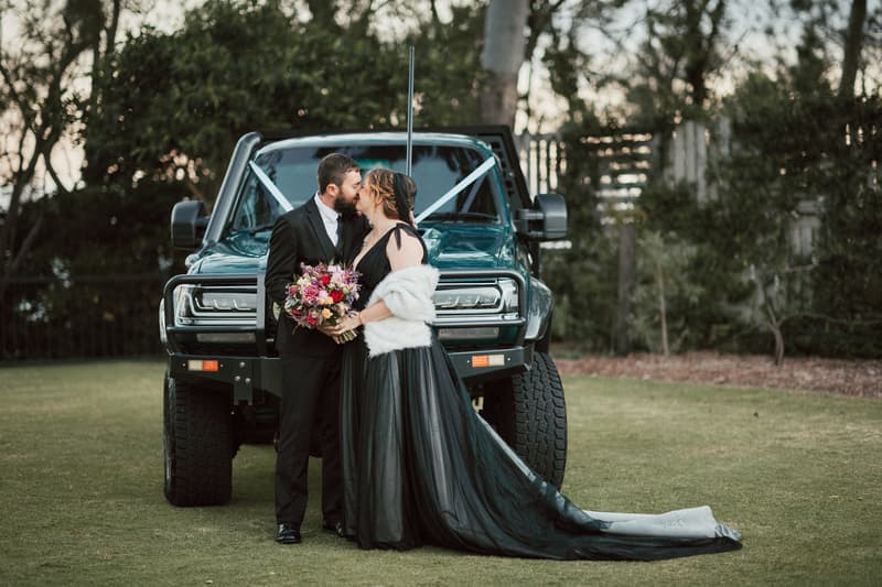Jayme and Christopher kiss in front of a large black vehicle outdoors, with Jayme holding a colorful bouquet and wearing a black gown with a white shawl.