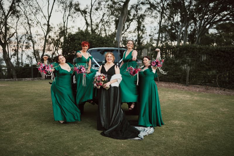 The bride Jayme in a black gown with a white shawl stands in front of a vehicle with four bridesmaids in green dresses holding floral hoops at Sandstone Point Hotel — The Pavilion.