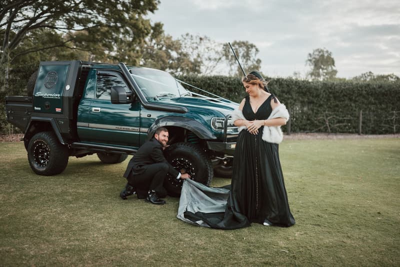 The groom crouches by a green four-wheel-drive vehicle adjusting the tire while the bride stands nearby in a black gown with a white shawl at Sandstone Point Hotel — The Pavilion.