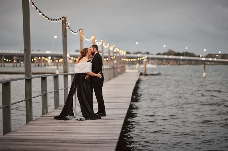 The bride and groom share a kiss on a wooden pier decorated with string lights at Sandstone Point Hotel.