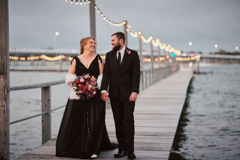 Jayme and Christopher walk hand in hand on a wooden pier with string lights overhead. Wedding photography coverage options by She Said Yes.