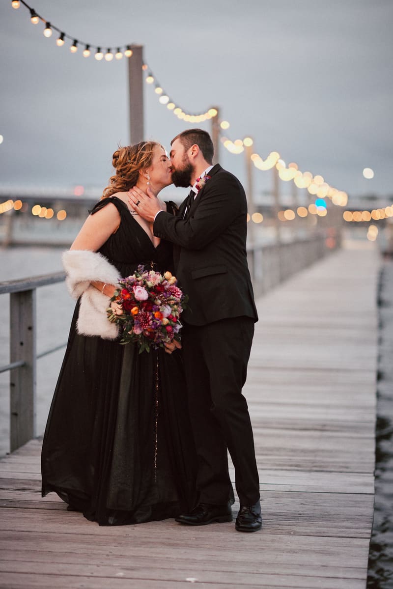 Jayme and Christopher share a kiss on a wooden pier decorated with string lights in the evening. Jayme wears a black gown and holds a colorful bouquet, while Christopher is dressed in a black suit with a boutonniere. Sandstone point hotel wedding by She Said Yes.