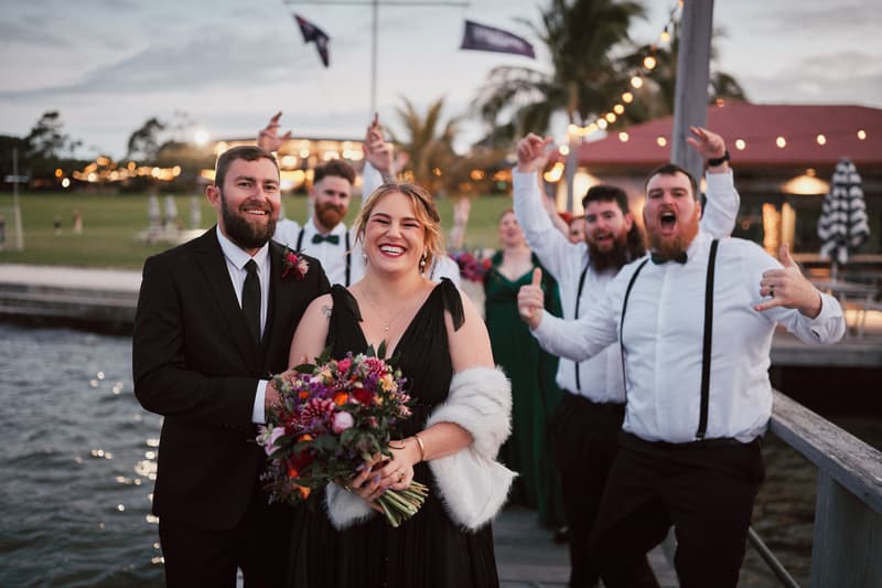 Christopher in a black suit and tie stands next to a bridesmaid in a black dress holding a bouquet at Sandstone Point Hotel — The Pavilion, with groomsmen and bridesmaids cheering behind them on a dock by the water.