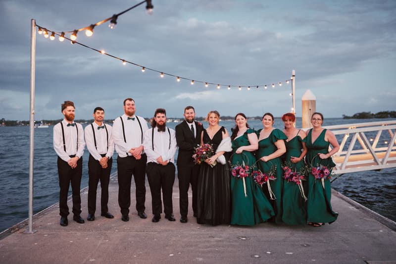 Jayme and Christopher pose with their bridal party on a pier at Sandstone Point Hotel — The Pavilion, with string lights overhead and water in the background.