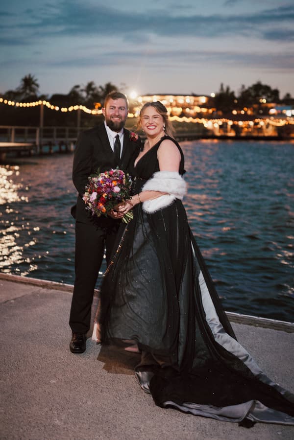 Jayme and Christopher pose together near water at dusk with string lights and a lit building in the background at Sandstone Point Hotel.
