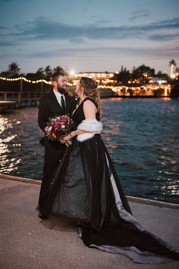 Jayme in a black wedding gown and white shawl holds a bouquet and faces Christopher in a black suit near the water at dusk with string lights and a lit building in the background at Sandstone Point Hotel.
