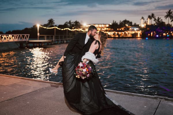 Jayme and Christopher share a kiss on a waterfront pier at dusk with string lights and a lit building in the background. Getting ready photos by She Said Yes.